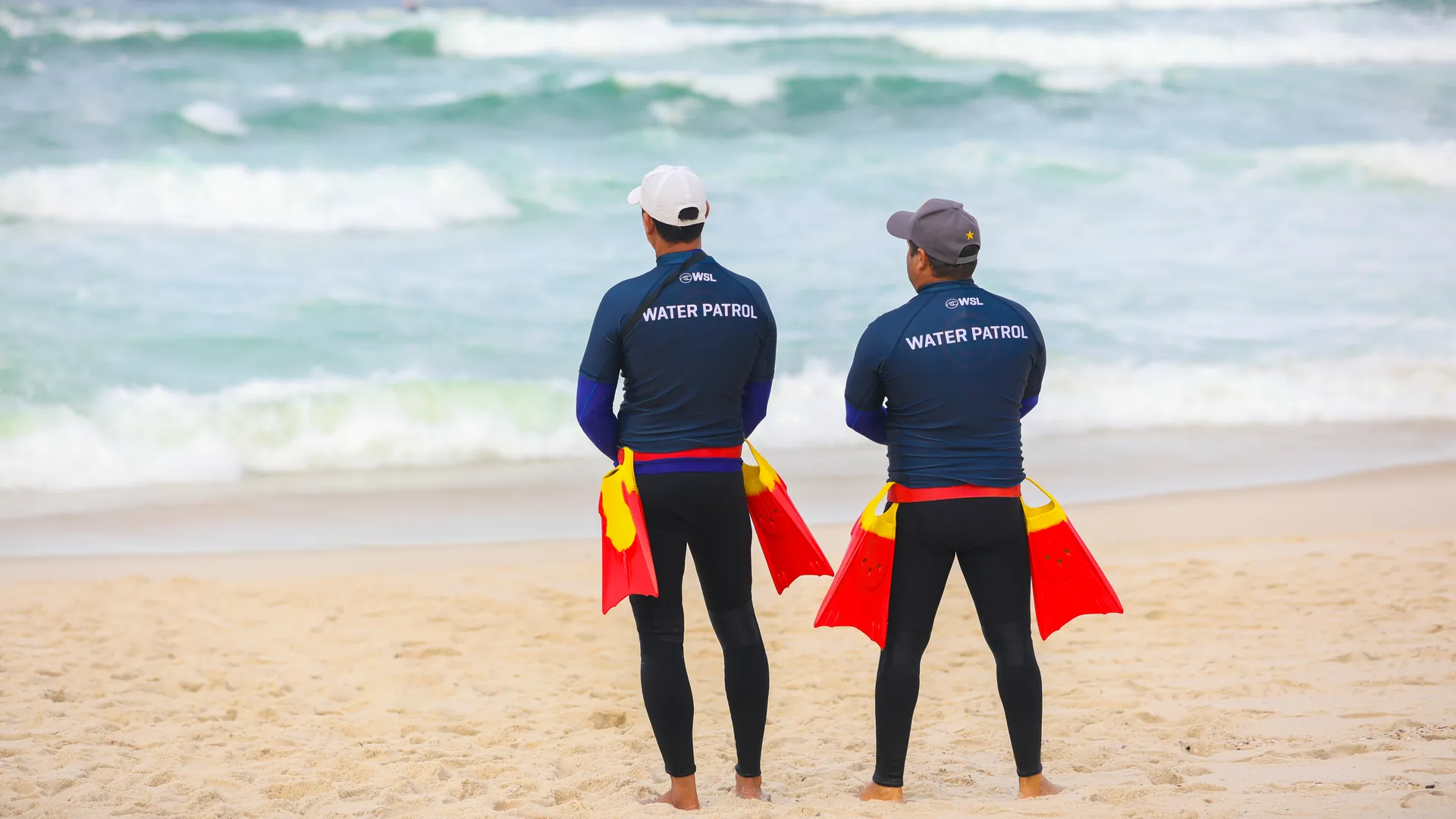 Dupla de resgate aquático na areia, de frente para o mar, com as nadadeiras na cintura